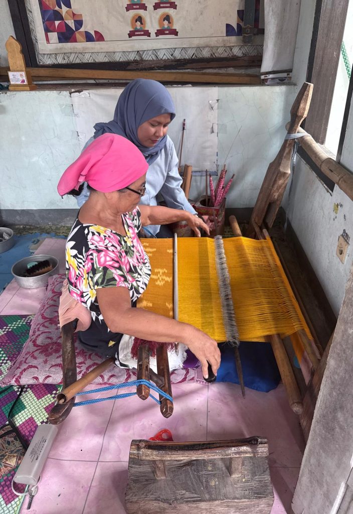 Mastawan sits with a lady who is hand weaving on a loom on the floor inside.